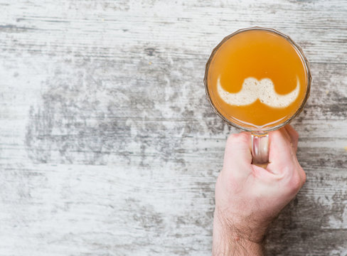 Man's Hand Holds A Mug Of Beer With Silhouettes Of Mustache On A Beer Foam. Top View. Space For Text