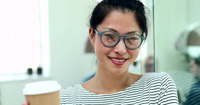 Young Asian businesswoman wearing glasses and smiling while standing in an office drinking a coffee with colleagues at work in the background