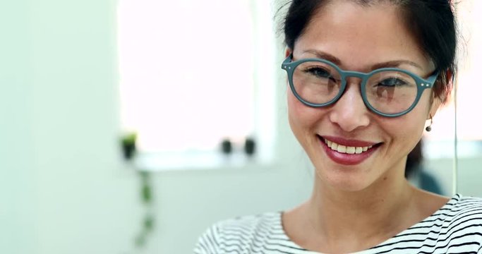 Smiling young Asian businesswoman wearing glasses standing with her arms crossed in a modern office with colleagues in the background