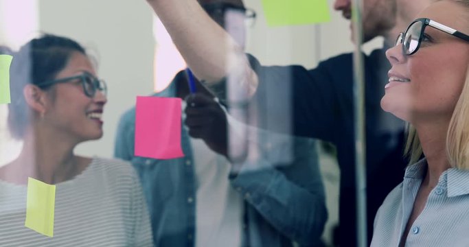 Diverse group of young business colleagues standing together in a modern office brainstorming with sticky notes on a glass wall