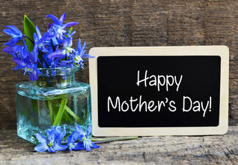 Happy Mother's Day greeting card with blue spring flowers un a glass vase and chalkboard on old wooden background.
Scilla siberica bouquet as a gift for mom.
Selective focus.