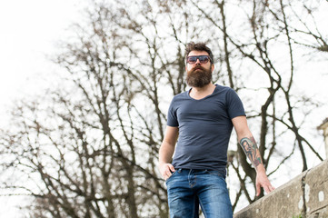 Naklejka na ściany i meble Barbershop and style concept. Man with beard on strict face, trees on background, defocused. Hipster with beard looks confident while standing outdoors. Bearded man wears modern sunglasses.