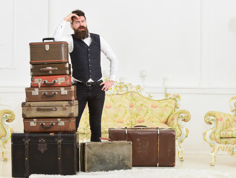 Baggage and relocation concept. Man with beard and mustache packed luggage, white interior background. Macho elegant on tired face, exhausted at end of packing, leans on pile of vintage suitcases.