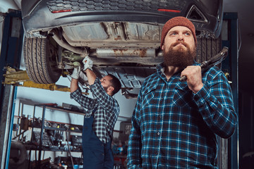 Two bearded brutal mechanics repair a car on a lift in the garage. 