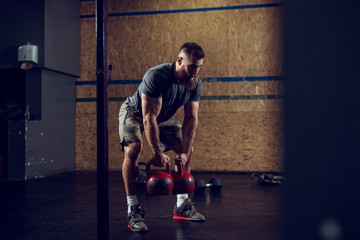 Portrait view of young bearded focused strong muscular shape bodybuilder man crouching with heavy kettlebells in hands at the gym.