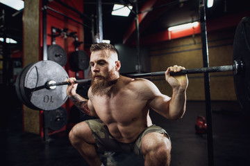 Fototapeta Portrait view of young bearded focused strong muscular fitness man crouching with a heavy barbell behind the neck while doing squats in the gym. obraz