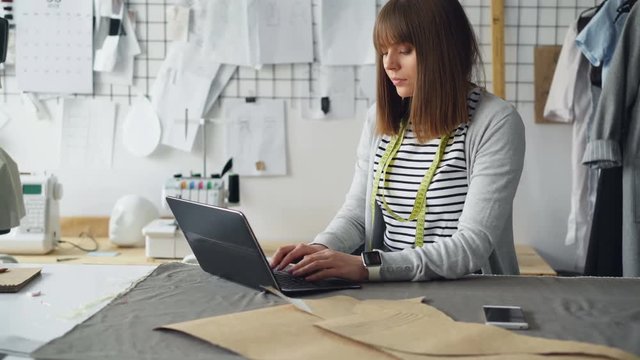 Female clothing design entrepreneur is using laptop to communicate with her clients and sell manufactured garments. Modern e-commerce in small business concept.