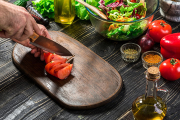 Man's hand cuts ripe red tomatoes for summer healthy vegetable salad on a wooden board, vertical