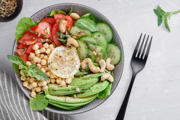 Top view of a healthy vegan lunch bowl with raw avocado, chickpeas, cashew, tomatoes, cucumber, greens and seeds over white kitchen table.