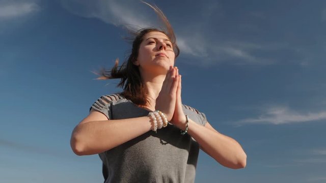 Young cheerful woman doing yoga asana on blue sky background. Girl in comfortable grey clothing meditating with namaste. Windy sunny weather. Health and grateful concept.