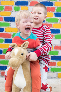 Two funny Caucasian children in bright clothes jumping on rocking horse against backdrop of multi-colored brick wall. The brothers are happy to play together. Concept of happy family and childhood
