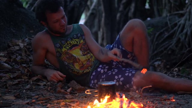 Caucasian Man Burning Little Campfire in the Tropical Forest at Twilight to Boil Kettle with Rice for Dinner.