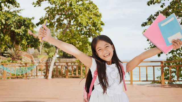 A charming philippine schoolgirl with a backpack and books in a park off the coast. A girl joyfully poses, raising her hands up with textbooks in her hands. Warm sunny day.