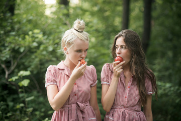 Camping in woods. Beautiful young girls wandering in forest. Two sisters in vintage dresses eating juicy tomatoes. Vitamins, nutrition and healthy diet concept