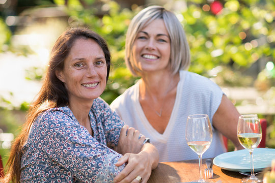  Portrait of two beautiful women in their forties. They are posing for the photo sits on a terrace in front of a glass of wine.