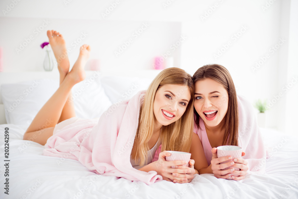 Wall mural Portrait of pretty, charming, cute, cheerful, attractive, successful girls in white and pink outfit having mugs with tea in hands, looking at camera, lying on stomach under plaid, blinking with eye