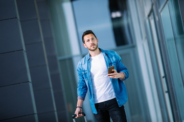 young businessman walking through the financial district of the city with his smartphone and suitcase. business concept