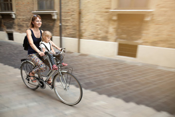 Fototapeta premium Mom with daughter riding bike in Italy. Child sitting in safety