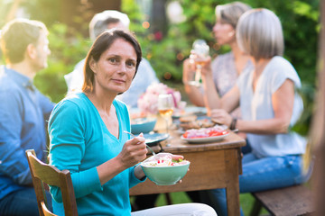 Looking at the camera a beautiful brunette woman in her forties with a bowl of mixed salad by hand. She sits around a table in a garden with friends for dinner.