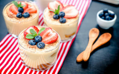 homemade, exquisite dessert tiramisu in glasses decorated with strawberry, blueberry, mint on black wooden table