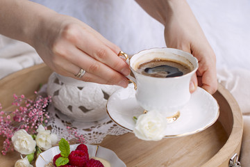 A woman is drinking coffee in bed. A wooden tray with breakfast. Raspberry berries and flowers. Light colors. Romance. Coffee mug in hands