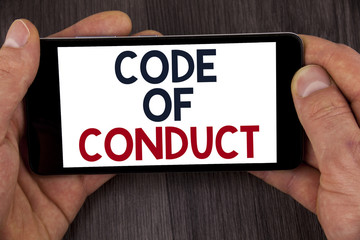 Writing note showing  Code Of Conduct. Business photo showcasing Follow principles and standards for business integrity written on Mobile Phone Screen holding by man on the wooden background.