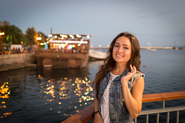 Pretty young woman walking on city promenade near sea in the evening. Soft colors. Street lights rays falls on her face. Happy girl enjoys vacation