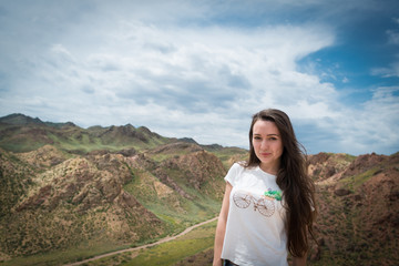 Young woman stands on background with view on the mountains and hills with long country road. Cloudy blue sky.
