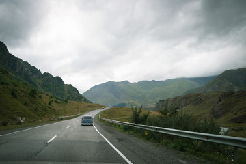 Small old stylish car is moving along the mountain highway. Scenic view on the road with majestic mountains on sides.