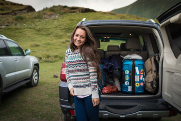 Young pretty woman traveler standing near open back door of the car with luggage.