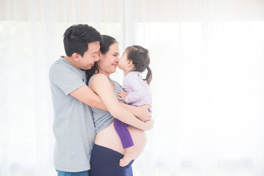 Little asian girl kissing her pregnant mother while her father hugging her mother from back