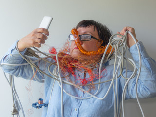 An elderly woman is tangled in wires. She looks at the smartphone. - Powered by Adobe