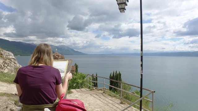 Young arthistic girl paints new art wit view of church of Saint John at Kaneo and Ohrid lake at background