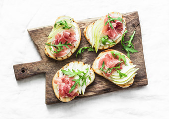 Bruschetta with cream cheese, pear, prosciutto and arugula on wooden chopping board on light...