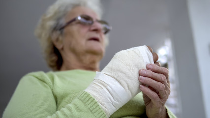 Sad old woman having pain sitting and holding her injured hand in bandages