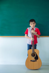 Young boy stading with guitar in front of blackboard in classroom