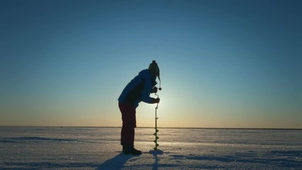 Man angler drilling hole in frozen lake for ice fishing on background sun in winter sky