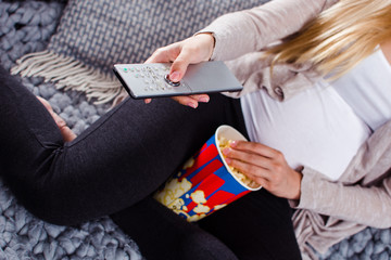 TV time! Close-up part of young pregnant woman watching TV and eating popcorn while sitting on the sofa in home interior