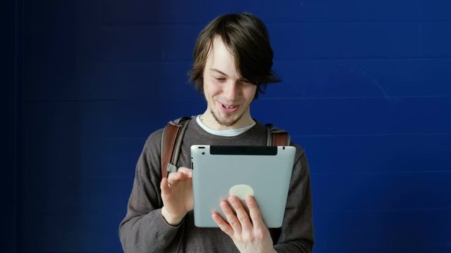 Student man uses a tablet for video communication and chatting with friends on a blue wall background