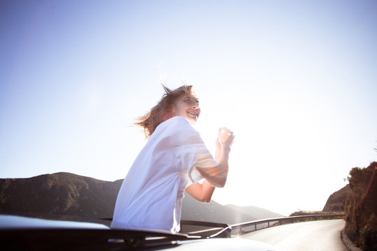 Happy excited beautiful young woman, free and careless teenager living the american dream on sunny empty road, stands up in convertible cabriolet car, wind in her hair, she laughs and smiles