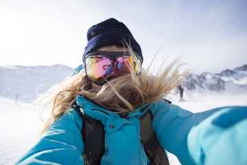 Cool, beautiful, young and attractive teenager or active lifestyle woman makes photo selfie on smartphone or action camera on ski slopes when snowboarding in mountains, wears goggles for snow and sun