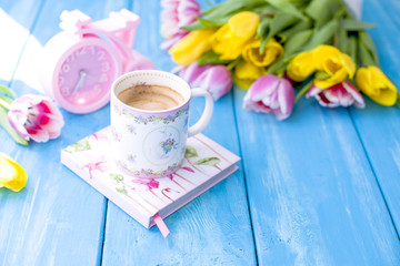Fototapeta na wymiar A cup of coffee on a blue wooden background. Bouquet of flowers yellow and pink. The pink clock is like a bicycle. Spring morning. Text