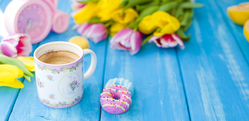 Fototapeta na wymiar A cup of coffee and donuts on a blue wooden background. Bouquet of flowers yellow and pink. The pink clock is like a bicycle. Spring morning. Text