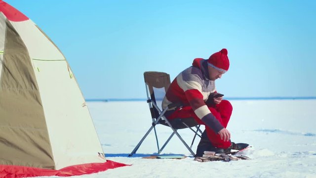 Fisherman sitting on chair at ice lake and using mobile phone on winter fishing