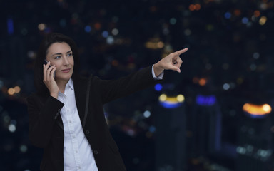 Caucasian businesswoman talking on her cell phone and pointing or touching touchscreen over blur colorful night light city tower, Business communication concept