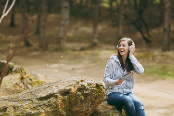 Young smiling woman in casual clothes sitting on stone listening music with headphones and tablet pc computer in city park or forest on green blurred background. Student lifestyle, leisure concept.