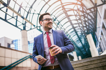 Businessman using mobile phone outside of office buildings in the background. Young caucasian man holding smartphone for business work and drink coffee to go.