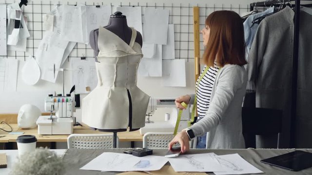 Self-employed female tailor is checking clothing patterns pinned to tailoring dummy then taking photos of it with smart phone. Nice workshop with sewing machines in background.