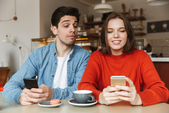 Image of happy woman holding mobile phone in cafe, while curious man looking at her smartphone with interest