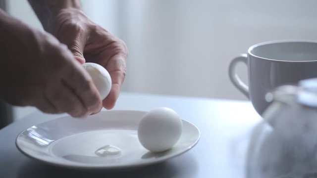 Man peels a hard boiled egg with hands in the morning. He stays near the table, there are cup and coffee pot